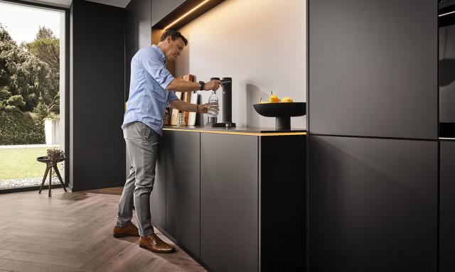 czarne fronty kuchenne, Pan nalewający wodę do butelki, patera na owoce, czarna bateria zlewozmywakowa, duże okno tarasowe, jodełka na podłodze, stołek, kwiat w doniczce, oświetlenie LED