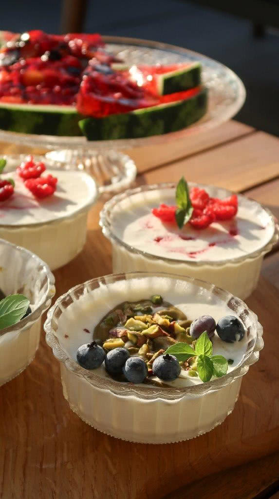 Domowe lody w szklanych pucharkach z pistacjami, borówkami i malinami, podane na drewnianym stole w letniej aranżacji.
