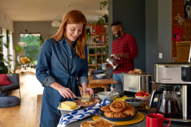 ekspres do kawy, toster, czajnik elektryczny, kobieta, smaruje pieczywo masłem, mężczyzna nalewa wodę do kubka, drewniana podłoga, szary blat kuchenny, duży stół z krzesłami