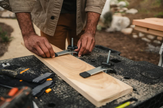 Mężczyzna mocuje drewnianą deskę za pomocą zacisków na składanym stole warsztatowym Keter Folding Work Table 2.0.