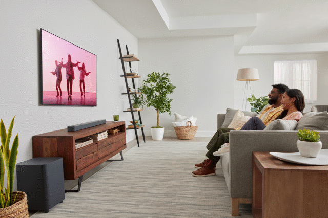 JBL Bar 500, półki, komoda, duży telewizor, kwiaty w doniczce, kanapa, dywan, duży głośnik, lampa podłogowa