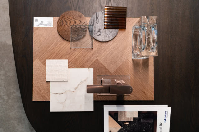 Moodboard wnętrzarski z próbkami drewna, kamienia i szkła oraz brązową baterią na tle drewnianej jodełki.