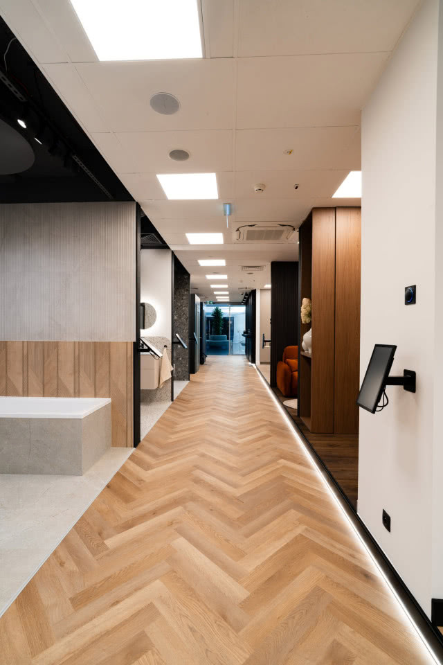 Nowoczesny showroom z drewnianą podłogą w jodełkę, ekspozycją łazienek i podświetlonym ciągiem komunikacyjnym.