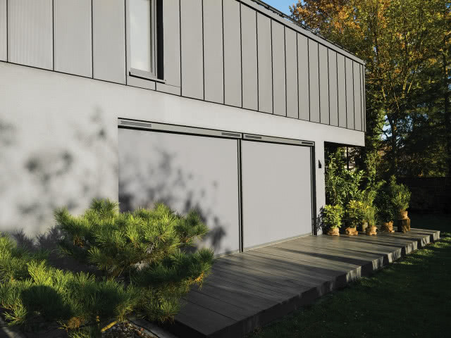 drewniany taras, markizy zewnętrzne, szara elewacja na budynku, iglaki, rośliny zielone w doniczkach, wysokie drzewa