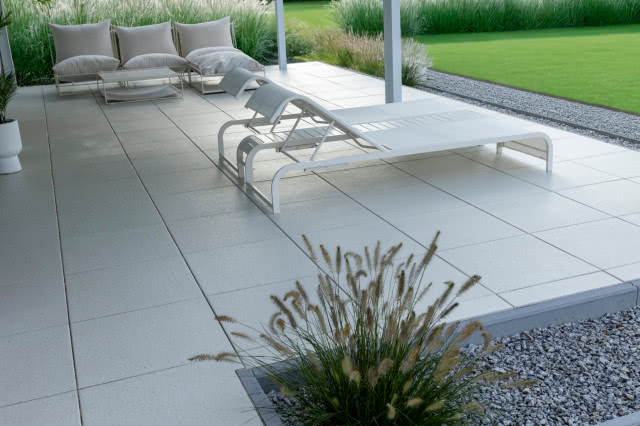 Nowoczesny taras z jasnych płyt, z leżakami i strefą wypoczynkową pod zadaszeniem, otoczony zielonym ogrodem i ozdobnymi trawami.