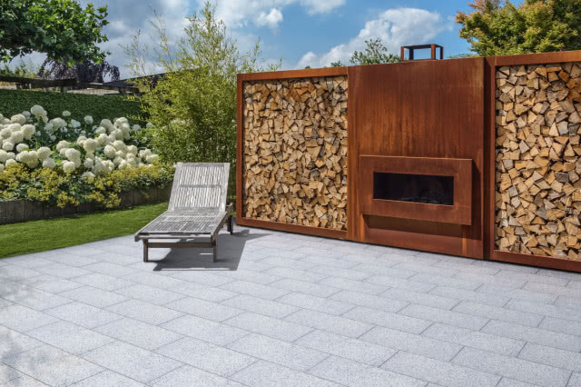 Taras z jasnych płyt Vestone z nowoczesnym kominkiem ogrodowym i ścianą na drewno