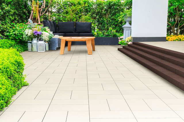 aras z jasnych płyt Vestone przy domu, z drewnianym stolikiem, sofą i schodami, otoczony bujną zielenią ogrodu.