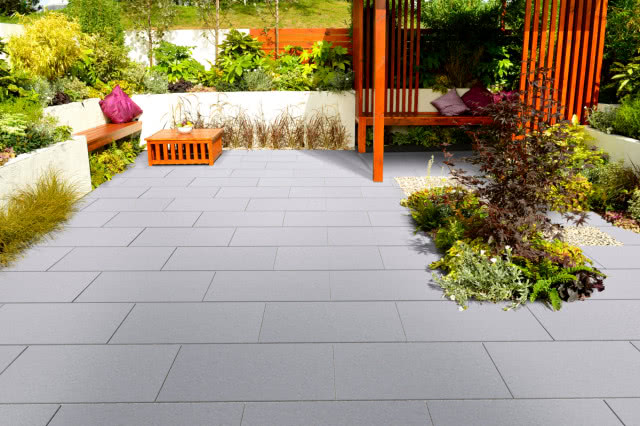 Nowoczesny taras z dużych, szarych płyt betonowych, z drewnianą zabudową wypoczynkową, ławką i stolikiem oraz bujną, wielopoziomową zielenią w podwyższonych rabatach.