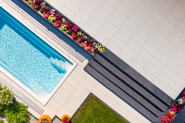 Nowoczesny taras z jasnych płyt w dużym formacie przy basenie, z ciemnymi schodami oraz długą rabatą kwiatową pełną kolorowych roślin, widok z góry.