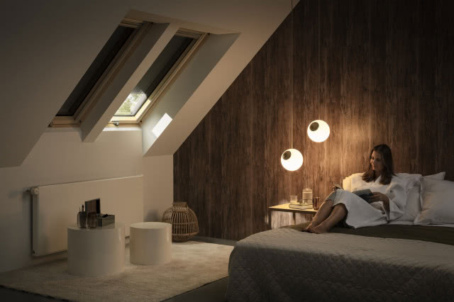 Solarna markiza zaciemniająca VELUX, sypialnia, duże łóżko, Kobieta leży na łóżku i czyta książkę, okna dachowe, stolik nocny, grzejnik