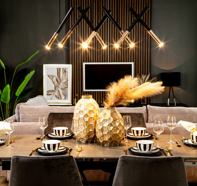 lamele na ścianie, duży stół złote wazony w kształcie ananasa, pióra, filiżanki, kieliszki, krzesła, telewizor, obrazy, kanapa, oświetlenie sufitowe