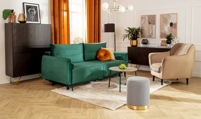 zielona kanapa, sztukateria na ścianie, komody, fotel, pufa, stolik kawowy, jabłka w misie zrobionej z drutu, kwiaty, wazony, obrazy, lampa podłogowa, oświetlenie sufitowe