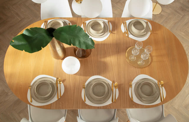 owalny stół drewniany, zastawa stołowa, taca, kieliszki, wazon z zielonymi liśćmi, sztućce, krzesła
