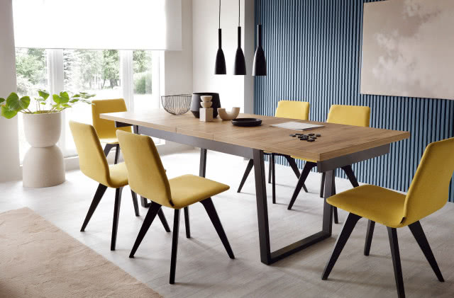 żółte krzesła na czarnych nogach, duży prostokątny stół, niebieskie panele na ścianie, trzy czarne wiszące nad stołem lampy, kwiat, białą roleta