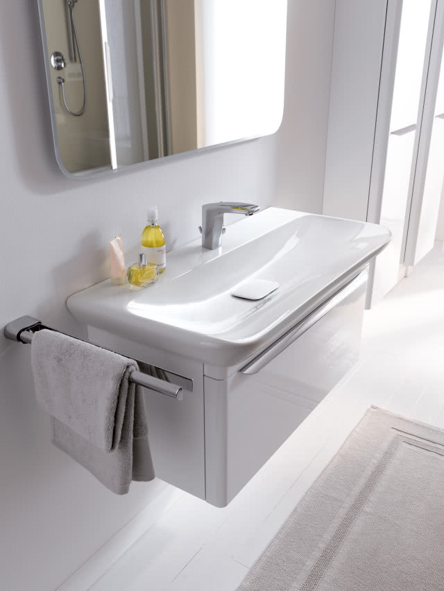 prostokątna umywalka z zaokrągleniami, wieszak na ręczniki, ręcznik, bateria umywalkowa, duże lustro, białe meble łazienkowe, dywanik