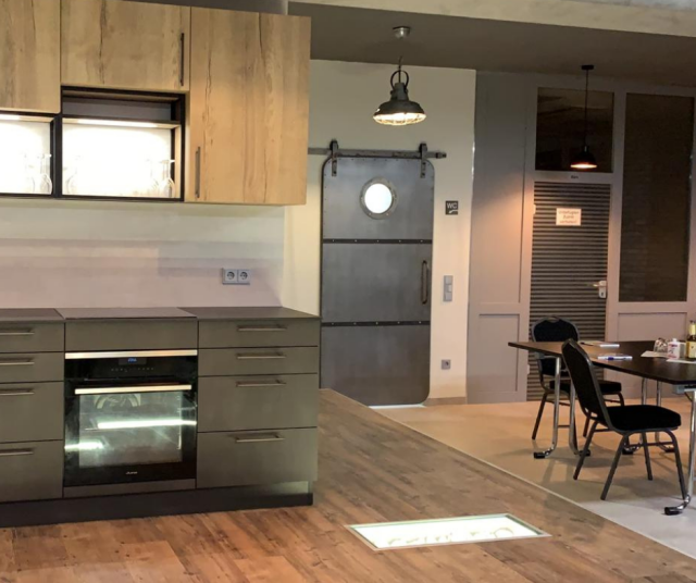 drzwi, szare fronty kuchenne dolnych szafek kuchennych, drewniane fronty kuchenne szafek wiszących, piekarnik, duży stół z krzesłami, drewniana podłoga