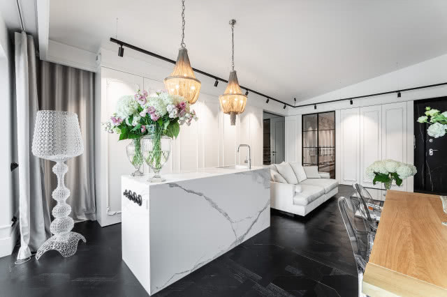 wyspa kuchenna, lampa podłogowa cała ze szkła, drewniany stół, krzesła, kanapa, wazony z kwiatami, zlewozmywak jednokomorowy, srebrna bateria zlewozmywakowa, granit, zasłony