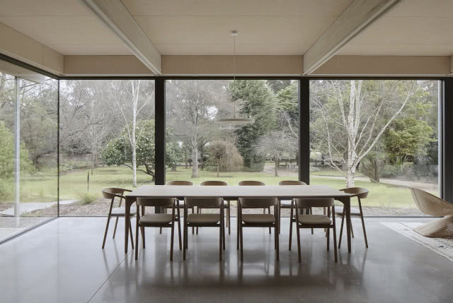 Długi drewniany stół na tle przeszklonej ściany i ogrodu.