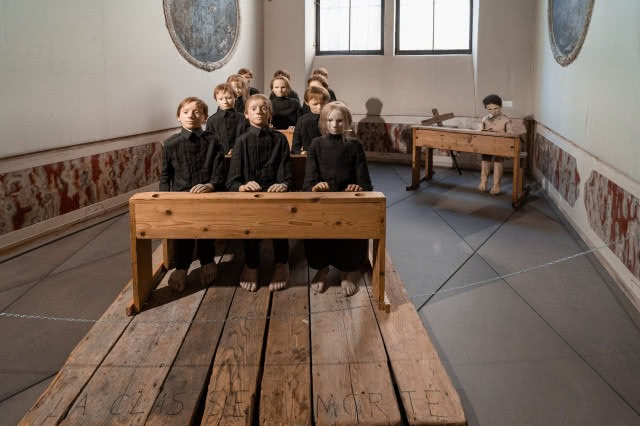 Instalacja Tadeusza Kantora: manekiny dzieci w szkolnej klasie siedzą przy drewnianych ławkach, tworząc niepokojącą scenę teatralną.