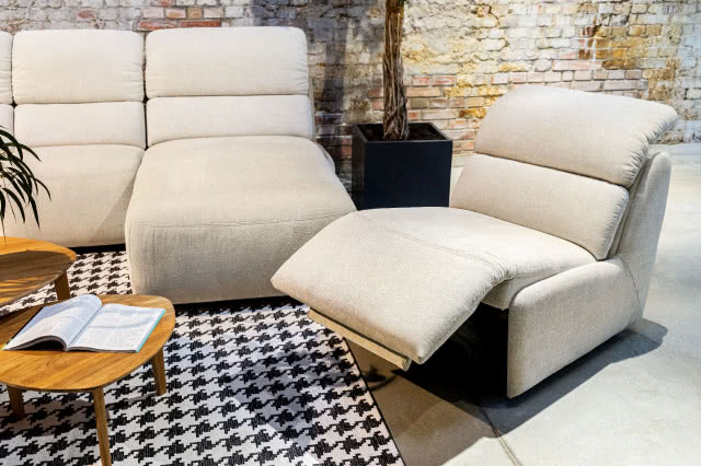 beżowa kanapa z rozkładanym siedziskiem, drewniany stolik kawowy, czarna donica, cegła na ścianie, książka, dywan biało czarny