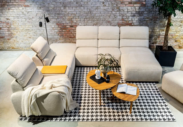 beżowa kanapa z rozkładanym siedziskiem, drewniany stolik kawowy, czarna donica, cegła na ścianie, książka, dywan biało czarny, czarna lampa podłogowa, rzeźba, wazony, koc