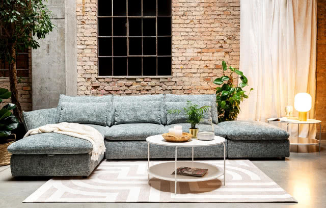 szara kanapa modułowa, cegła na ścianie, biało beżowy dywan, okrągły stolik kawowy, czasopisma, świece, kwiat, pled