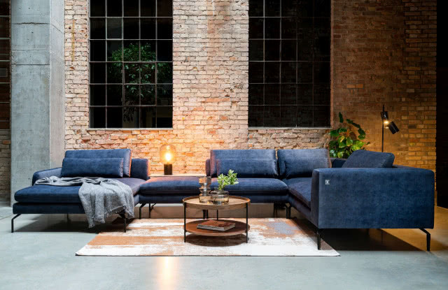 granatowa kanapa, okrągły stolik kawowy, wazon, kwiaty, dywan, na ścianie cegła, czarna lampa podłogowa, lampa stojąca na szafce 