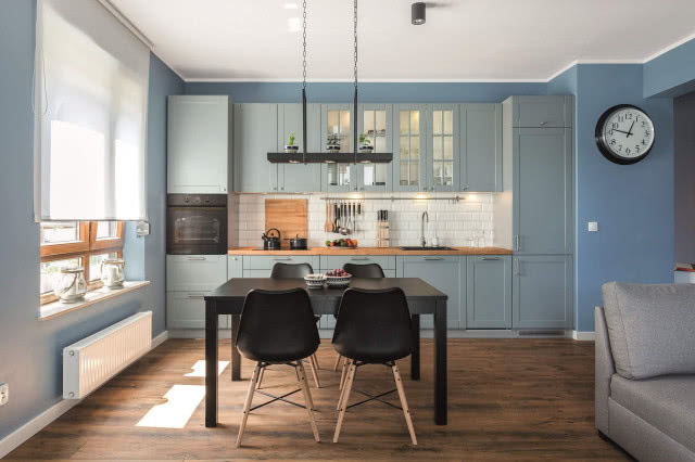 niebieskie meble kuchenne, niebieskie ściany, czarny duży stół z krzesłami, zegar na ścianie, drewniany blat kuchenny, rolety, drewniana podłoga