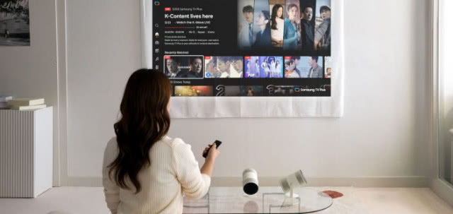 Kobieta steruje projektorem Samsung The Freestyle, oglądając platformę streamingową wyświetlaną na ścianie w jasnym wnętrzu