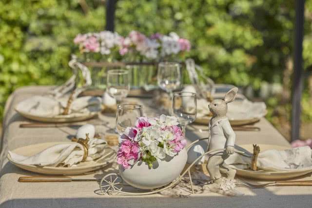 Figurka zająca pchającego taczkę pełną białych i różowych pelargonii