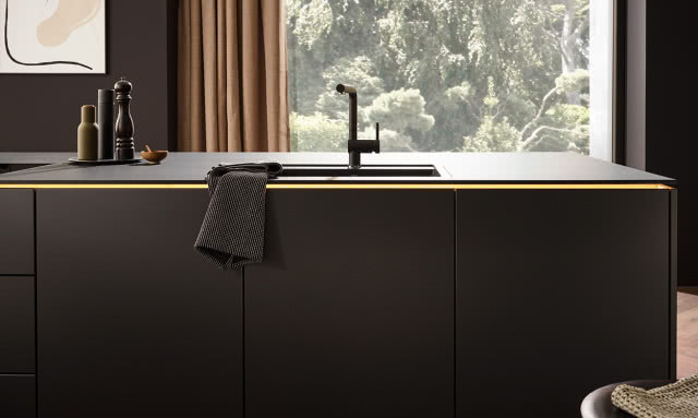 czarne, matowe fronty kuchenne, wyspa kuchenna z czarnym blatem, czarna płyta indukcyjna, czarny zlew, czarna bateria zlewozmywakowa, ekspres do kawy, wazon, misa na owoce, książka kucharska, stołek, piekarnik, przyprawy, drewniane deski do krojenia, moździerz 