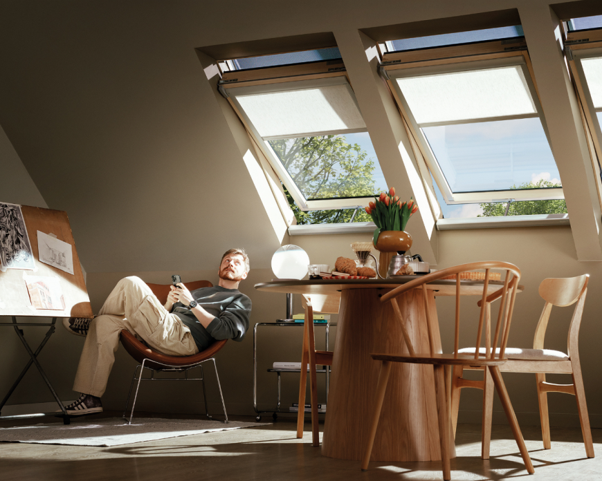 Mężczyzna relaksuje się na poddaszu przy oknach dachowych z roletami, jasne wnętrze z drewnianym stołem i naturalnym światłem