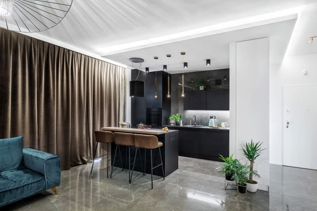 czarne meble kuchenne, czarne AGD, wyspa kuchenna, stołki przy wyspie kuchennej, kanapa, brązowe zasłony, oświetlenie sufitowe punktowe, kwiaty w doniczkach