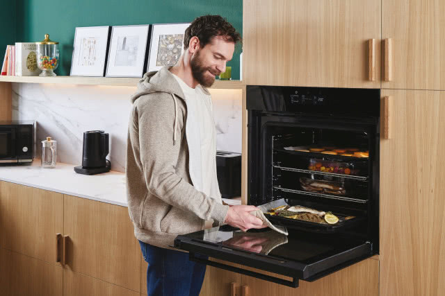 Mężczyzna wyjmujący blaszkę z jedzeniem z piekarnika, czajnik elektryczny, mikrofalówka, obrazy, półka