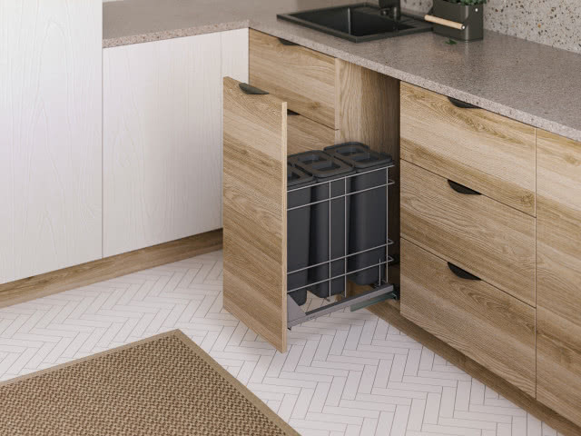 kosze do segregacji śmieci, drewniane fronty kuchenne, czarny zlewozmywak, na podłodze biała jodełka, beżowo-brązowy dywan, białe fronty kuchenne