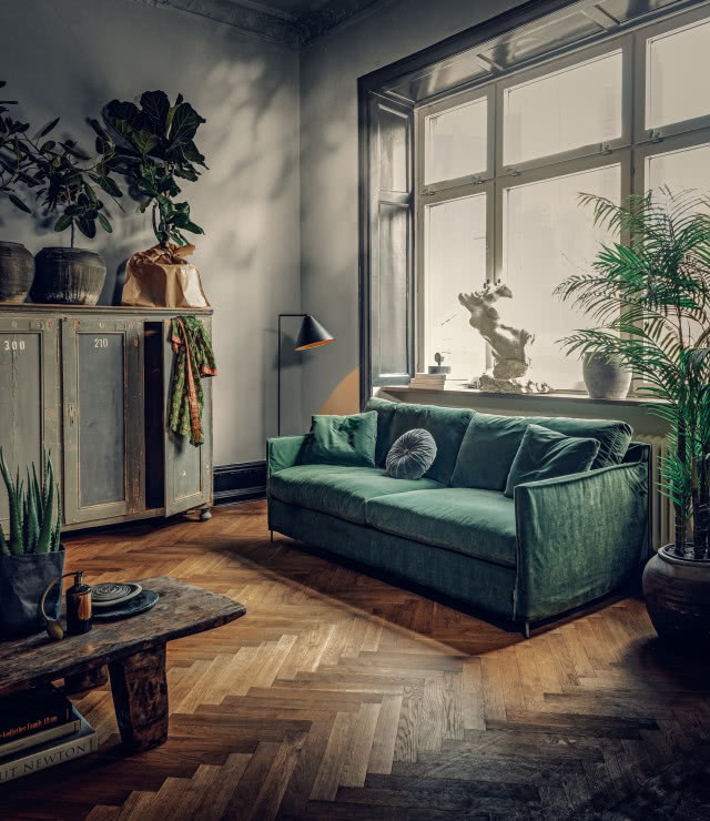 zielona sofa, komoda, stolik, książki, rzeźby, kwiaty zielone, jodełka na podłodze, lampa podłogowa