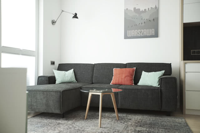 szary narożnik, poduszki ozdobne, obraz lampa ścienna, stolik kawowy, dywan, komoda