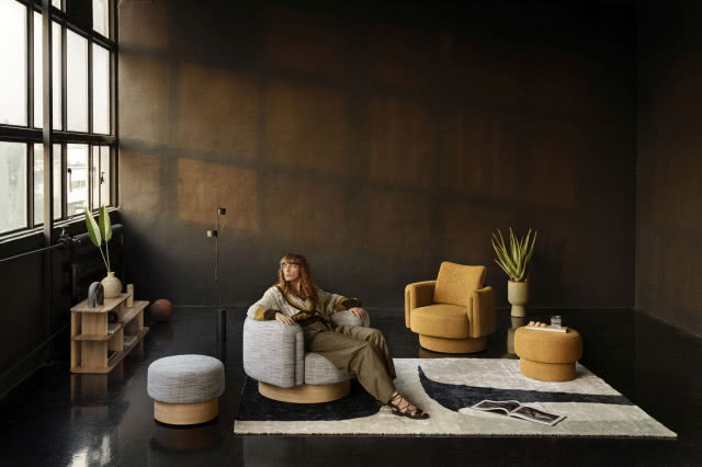 fotele, brązowe ściany, pufa, dywan, kobieta siedząca na fotelu, kwiaty, lampa podłogowa duże okno, grzejnik