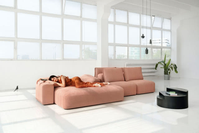 sofa kolorze pudrowego różu, pani śpiąca na sofie, stolik kawowy, kwiat w doniczce, oświetlenie sufitowe, duże okna, grzejnik