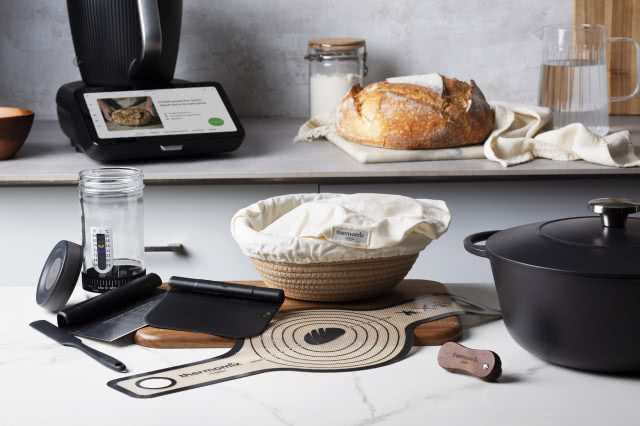 Zestaw Thermomix do pieczenia domowego chleba z koszykiem do wyrastania ciasta, naczyniem żeliwnym, akcesoriami i świeżym bochenkiem.