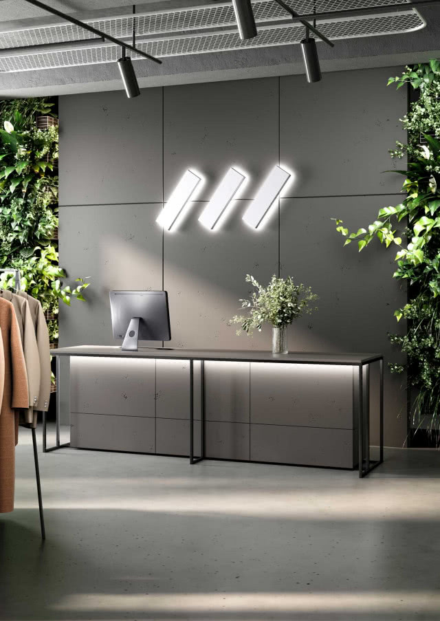 Nowoczesna lada recepcyjna z frontami RAUVISIO CUBE w odcieniu betonu, otoczona zielenią wertykalną i designerskim oświetleniem ściennym