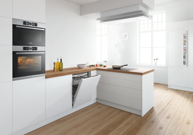 piekarnik, ekspres do kawy, zmywarka, płyta indukcyjna, wyspa kuchenna, białe meble kuchenne, drewniane blaty, duży stół z krzesłami, obrazki, oświetlenie sufitowe, drewniana podłoga, duże okno, patelnia