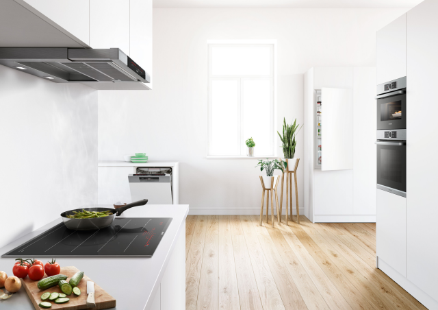 piekarnik, ekspres do kawy, zmywarka, płyta indukcyjna, wyspa kuchenna, białe meble kuchenne, drewniane blaty, duży stół z krzesłami, obrazki, oświetlenie sufitowe, drewniana podłoga, duże okno, patelnia, okap, lodówka, kwiaty w doniczce, warzywa, deska do krojenia, nóż