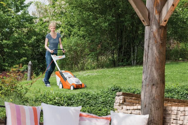Kompaktowa kosiarka elektryczna STIHL RME 339 C z wygodnym pojedynczym uchwytem, kobieta kosi trawę, ogród, drzewa, taras, poduszki ozdobne, belka, mur z cegieł