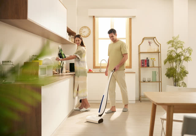 białe fronty kuchenne, drewniane blaty, stół z krzesłąmi, zegar, okno, mop bioniczny, Pan sprzątający mopem, Pani zmywa naczynia, kwiat, regał