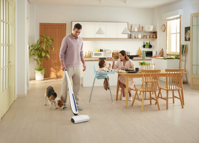 Tata sprzątający mopem bionicznym w kuchni, pies, mama karmi dziecko, stół z krzesłami. białe meble kuchenne, drewniane drzwi, duży kwiat zielony w doniczce