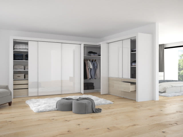 Bezuchwytowe otwieranie szafek, garderoba, ubrania, półki, szuflady, drzwi przesuwne, fotel, pufy, koc, dywanik, sypialnia, duże łóżko, duże okno, szare zasłony, jasna drewniana podłoga