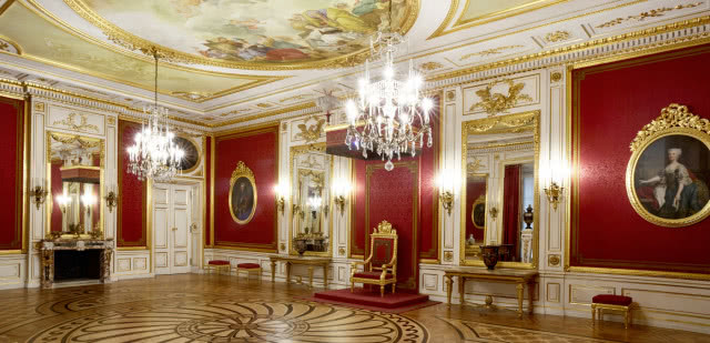 na tle ściany, obitą czerwoną tkaniną, stoi tron pod baldachimem, ujęcie całej sali