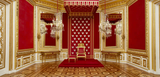 na tle ściany, obitą czerwoną tkaniną, stoi tron pod baldachimem