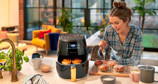 frytkownica beztłuszczowa, pączki, miseczki, kubek z kawą czajniczek do kawy, bateria zlewozmywakowa, kubki, fotel, duży stół, kwiaty w doniczce, duże okno